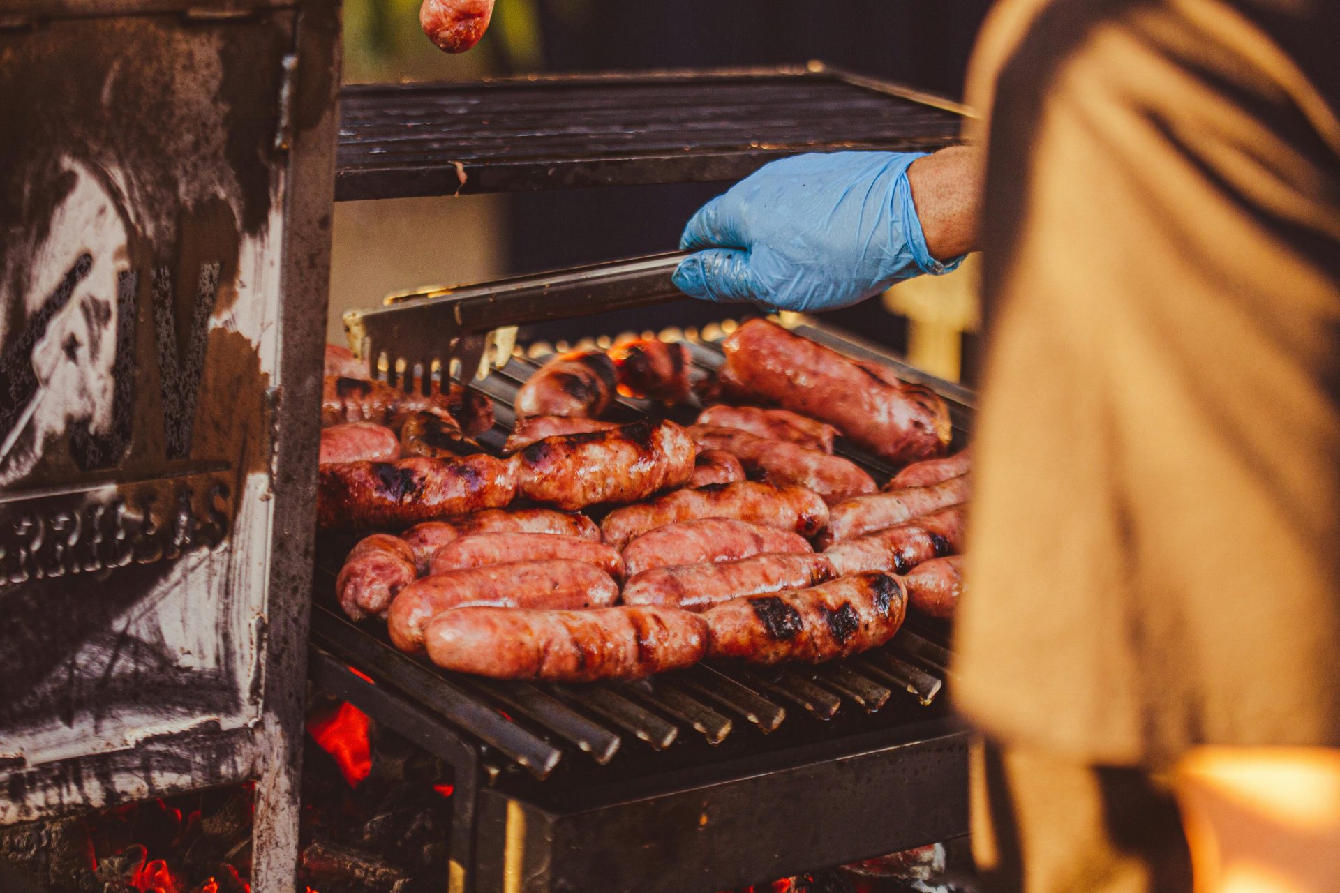 A hand with a glove uses tongs to turn sausages on an outdoor grill, flames visible.